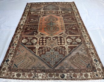 Puede incluir: Una alfombra rectangular con un medallón central naranja rodeado por un campo marrón oscuro con intrincados patrones geométricos y florales. El borde muestra un motivo repetitivo en crema, marrón y negro, acabado con flecos blancos.