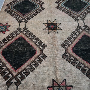 May include: A vintage rug with a cream background and a geometric pattern in black, brown, pink, and blue. The pattern features repeating diamond shapes with stars in the center.