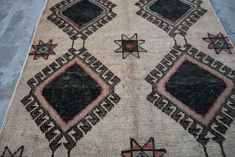 May include: A vintage rug with a cream background and a geometric pattern in black, brown, pink, and blue. The pattern features repeating diamond shapes with stars in the center.