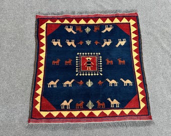 Puede incluir: Una alfombra azul con un borde rojo y un patrón geométrico amarillo y blanco. La alfombra presenta un diseño central de una figura estilizada con camellos y otros animales.