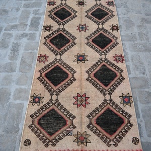 May include: A beige rug with a geometric pattern of black diamonds, red and blue stars, and brown and white accents. The rug has a fringe border.