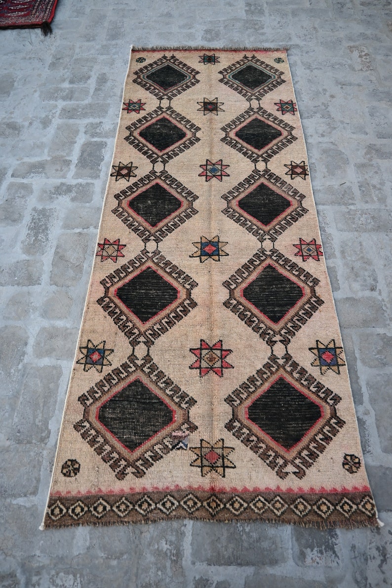 May include: A beige rug with a geometric pattern of black diamonds, red and blue stars, and brown and white accents. The rug has a fringe border.