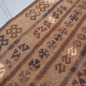 K&ouml;nnte beinhalten: Ein brauner und beigefarbener Teppich mit geometrischen Mustern. Der Teppich hat eine traditionelle gewebte Textur.