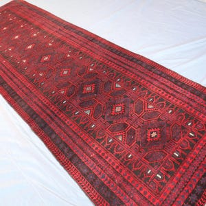 May include: A long, rectangular runner rug featuring a deep red and black color palette. The surface is decorated with a repeating geometric pattern of diamonds and intricate tribal motifs, finished with short white fringe at the ends.