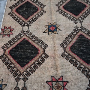May include: A vintage rug with a cream background and a geometric pattern. The pattern features black diamonds with red and brown borders, and star-shaped motifs in shades of red, blue, and yellow.