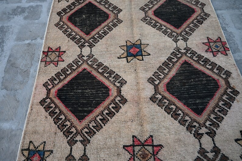 May include: A vintage rug with a cream background and a geometric pattern. The pattern features black diamonds with red and brown borders, and star-shaped motifs in shades of red, blue, and yellow.