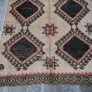 May include: A beige rug with a geometric pattern in black, red, and brown. The rug has a diamond shape with a star in the center of each diamond. The rug has a fringe border.