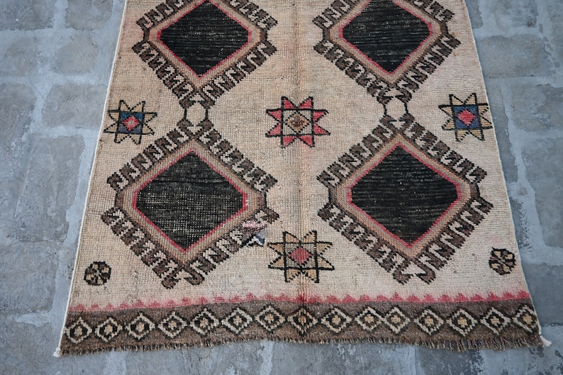 May include: A beige rug with a geometric pattern in black, red, and brown. The rug has a diamond shape with a star in the center of each diamond. The rug has a fringe border.
