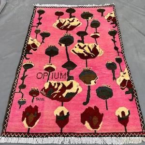 K&ouml;nnte beinhalten: Ein rechteckiger, pinkfarbener Teppich mit einem Blumenmuster mit gro&szlig;en, stilisierten Blumen in Creme, Burgunderrot und Schwarz. Das Wort "OPIUM" ist auf dem Teppich zu sehen. Der Teppich hat einen Fransenrand.