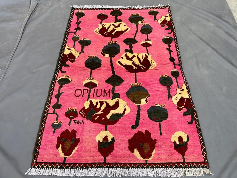 K&ouml;nnte beinhalten: Ein rechteckiger, pinkfarbener Teppich mit einem Blumenmuster mit gro&szlig;en, stilisierten Blumen in Creme, Burgunderrot und Schwarz. Das Wort "OPIUM" ist auf dem Teppich zu sehen. Der Teppich hat einen Fransenrand.