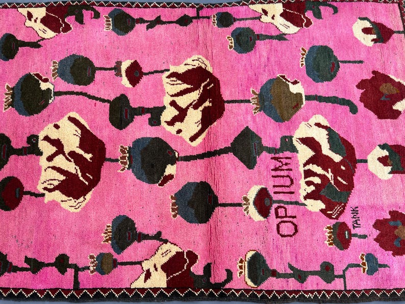 K&ouml;nnte beinhalten: Ein pinkfarbener Teppich mit einem sich wiederholenden Muster aus stilisierten Blumen und Samenkapseln in Wei&szlig;, Burgunderrot und Blaugr&uuml;n. Das Wort "OPIUM" ist vertikal in Schwarz gedruckt. Der Teppich hat einen dunklen Rand.