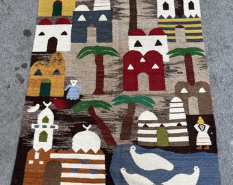 Puede incluir: Alfombra tejida a mano con una escena de pueblo con edificios, palmeras y pájaros. La alfombra tiene un fondo marrón, beige y azul con acentos de rojo, amarillo y blanco. La alfombra es rectangular con flecos en los extremos cortos.