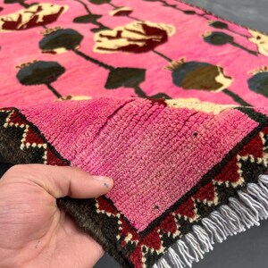K&ouml;nnte beinhalten: Ein pinkfarbener Teppich mit einem Blumenmuster in Gelb-, Braun- und Gr&uuml;nt&ouml;nen. Der Teppich hat einen dunkelbraunen Rand mit roten und wei&szlig;en geometrischen Mustern. Wei&szlig;e Fransen.