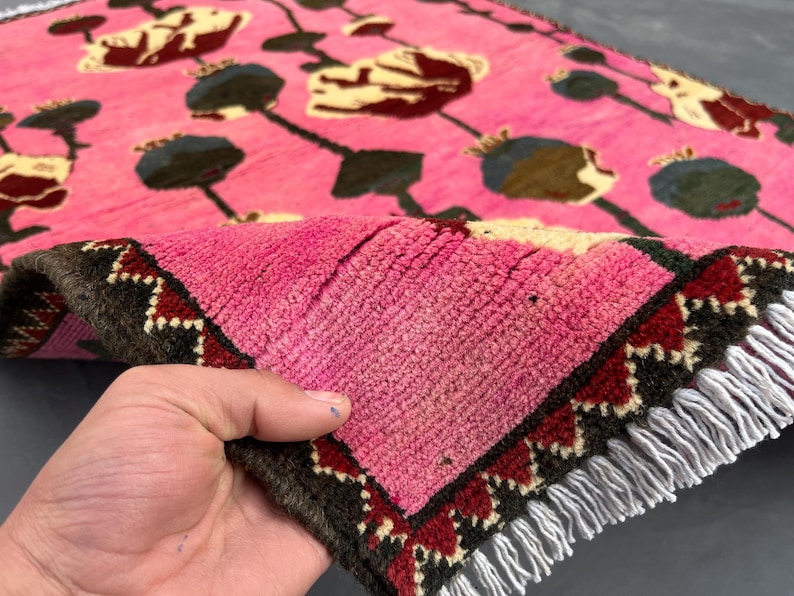 K&ouml;nnte beinhalten: Ein pinkfarbener Teppich mit einem Blumenmuster in Gelb-, Braun- und Gr&uuml;nt&ouml;nen. Der Teppich hat einen dunkelbraunen Rand mit roten und wei&szlig;en geometrischen Mustern. Wei&szlig;e Fransen.