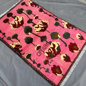 K&ouml;nnte beinhalten: Ein rechteckiger, rosa Teppich mit floralem Muster. Der Teppich zeigt ein sich wiederholendes Design aus stilisierten Blumen in Creme, Burgunderrot und Dunkelgr&uuml;n, mit schwarzen Stielen und Samenkapseln. Das Wort "OPIUM" ist sichtbar.