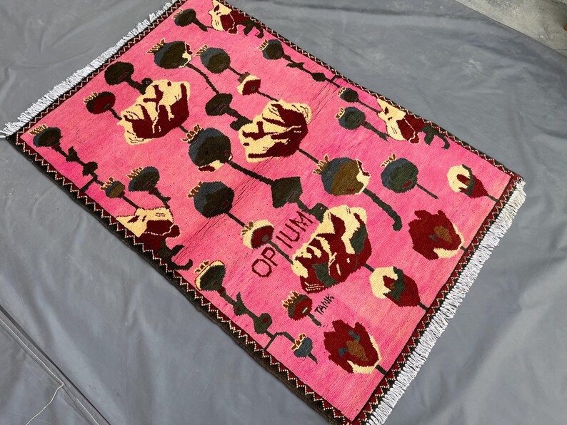 K&ouml;nnte beinhalten: Ein rechteckiger, rosa Teppich mit floralem Muster. Der Teppich zeigt ein sich wiederholendes Design aus stilisierten Blumen in Creme, Burgunderrot und Dunkelgr&uuml;n, mit schwarzen Stielen und Samenkapseln. Das Wort "OPIUM" ist sichtbar.