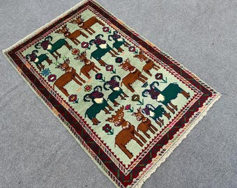 Puede incluir: Una alfombra verde claro con un borde rojo, negro y blanco. La alfombra presenta un patrón repetitivo de animales estilizados, incluyendo ciervos y carneros, en tonos de marrón, verde y negro.
