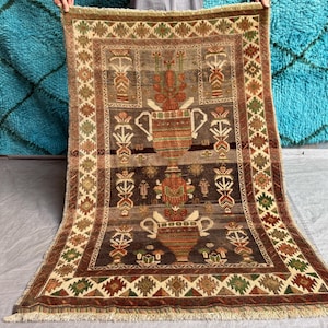 May include: A rectangular woven rug featuring a central design of stacked urns in shades of brown, orange, and green. The rug has a wide border with a repeating geometric diamond pattern and a fringe along the top and bottom edges.