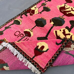 K&ouml;nnte beinhalten: Ein pinkfarbener Teppich mit einem sich wiederholenden Blumenmuster in Schwarz, Creme und Burgunder. Der Teppich hat einen dunkelbraunen Zickzack-Rand und wei&szlig;e Fransen. Das Wort "OP" ist auf der Oberfl&auml;che des Teppichs zu sehen.