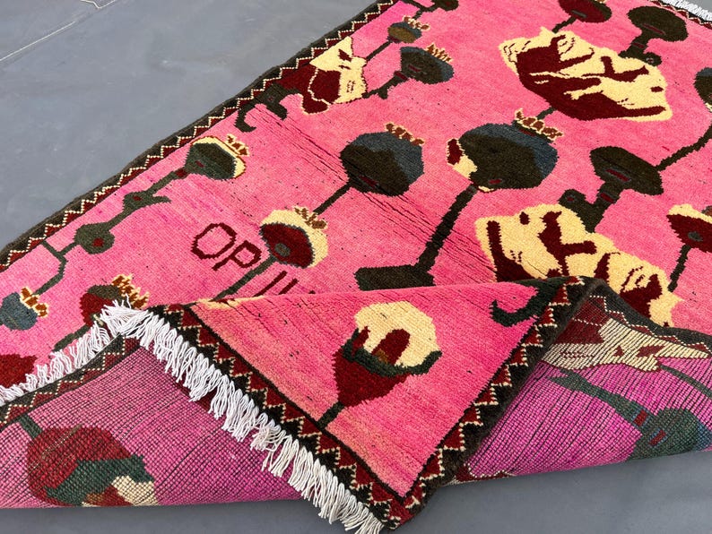 K&ouml;nnte beinhalten: Ein pinkfarbener Teppich mit einem sich wiederholenden Blumenmuster in Schwarz, Creme und Burgunder. Der Teppich hat einen dunkelbraunen Zickzack-Rand und wei&szlig;e Fransen. Das Wort "OP" ist auf der Oberfl&auml;che des Teppichs zu sehen.