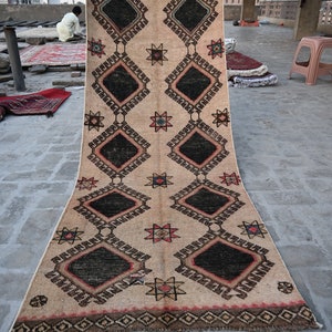 May include: A beige rug with a geometric pattern of black, red, and blue diamonds and stars. The rug is fringed on all sides.