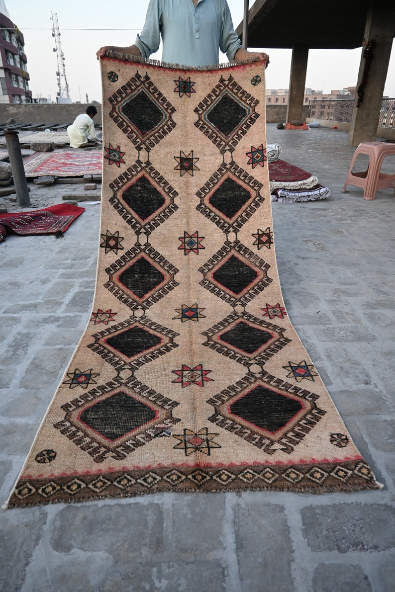 May include: A beige rug with a geometric pattern of black, red, and blue diamonds and stars. The rug is fringed on all sides.