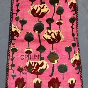 K&ouml;nnte beinhalten: Ein rechteckiger, rosa Teppich mit floralem Muster. Der Teppich zeigt stilisierte Mohnblumen in Creme, Burgunderrot und Dunkelgr&uuml;n, mit schwarzen Stielen und Samenkapseln. Das Wort "OPIUM" ist in der Mitte zu sehen, darunter "TANK".