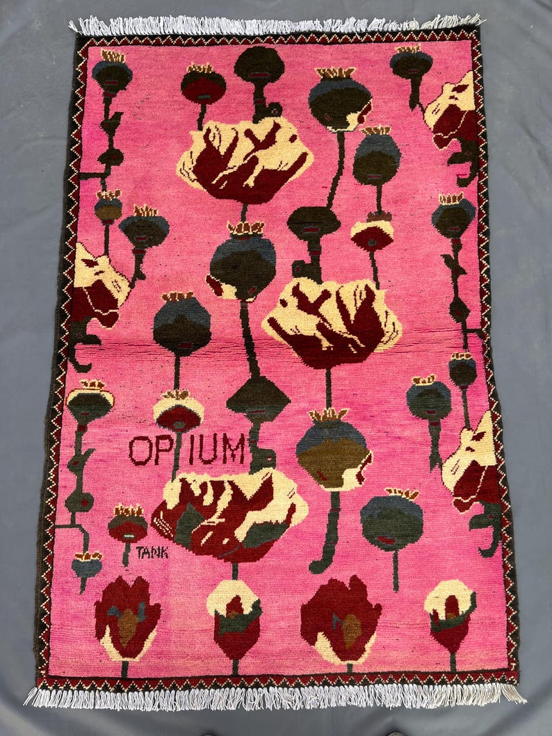 K&ouml;nnte beinhalten: Ein rechteckiger, rosa Teppich mit floralem Muster. Der Teppich zeigt stilisierte Mohnblumen in Creme, Burgunderrot und Dunkelgr&uuml;n, mit schwarzen Stielen und Samenkapseln. Das Wort "OPIUM" ist in der Mitte zu sehen, darunter "TANK".