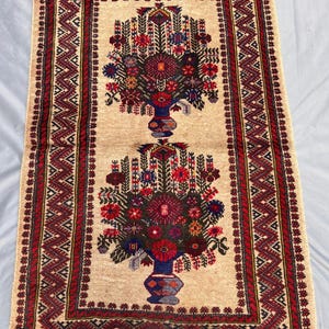 May include: Rectangular beige rug with a central floral design in red, blue, and green. The rug features a border with geometric patterns in red and dark brown, and fringed edges. The floral design is in a vase.