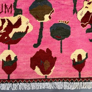 K&ouml;nnte beinhalten: Ein pinkfarbener Teppich mit einem sich wiederholenden Blumenmuster in Rot-, Creme- und Gr&uuml;nt&ouml;nen. Das Wort "OPIUM" ist in der oberen linken Ecke sichtbar. Der Teppich hat einen Fransenrand.