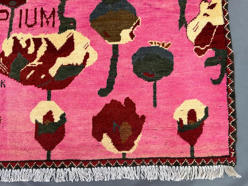 K&ouml;nnte beinhalten: Ein pinkfarbener Teppich mit einem sich wiederholenden Blumenmuster in Rot-, Creme- und Gr&uuml;nt&ouml;nen. Das Wort "OPIUM" ist in der oberen linken Ecke sichtbar. Der Teppich hat einen Fransenrand.