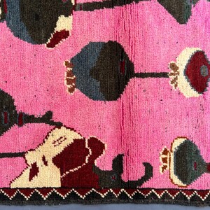 K&ouml;nnte beinhalten: Ein pinkfarbener Teppich mit einem sich wiederholenden Muster aus stilisierten Blumenformen in Dunkelgrau, Burgunderrot und Creme. Das Wort "OPIUM" ist vertikal in Burgunderrot dargestellt. Der Teppich hat einen dunkelgrauen Rand.