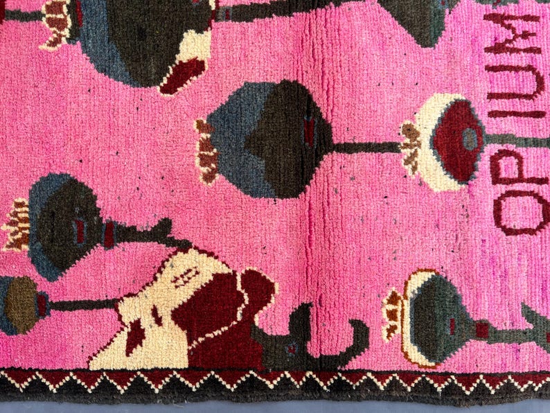 K&ouml;nnte beinhalten: Ein pinkfarbener Teppich mit einem sich wiederholenden Muster aus stilisierten Blumenformen in Dunkelgrau, Burgunderrot und Creme. Das Wort "OPIUM" ist vertikal in Burgunderrot dargestellt. Der Teppich hat einen dunkelgrauen Rand.