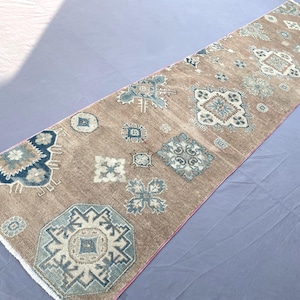 May include: A long, rectangular runner rug featuring a light brown base color with a repeating pattern of geometric shapes and floral motifs in shades of blue, white, and dark navy. The rug has a low-pile texture and finished edges on both ends.