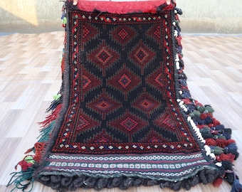 May include: A black and red rug with a geometric pattern and fringe. The rug has a traditional design with a central diamond shape and smaller diamonds surrounding it.
