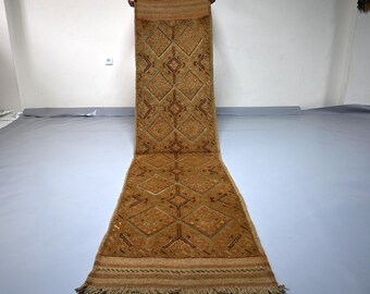 May include: A long, rectangular runner rug in shades of brown and tan, featuring a repeating pattern of diamonds with dark brown geometric accents. The rug has a textured pile, a fringed edge at the bottom, and a horizontal striped border design.