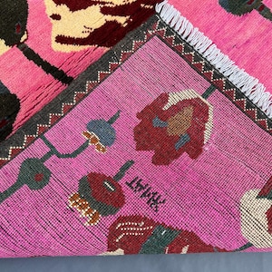 K&ouml;nnte beinhalten: Ein leuchtend rosa Teppich mit einem Blumenmuster in Dunkelgr&uuml;n, Creme und Burgunderrot. Der Teppich hat einen dunkelgrauen Rand mit Zickzackmuster und wei&szlig;en Fransen. Das Design zeigt stilisierte Blumen und geometrische Formen.