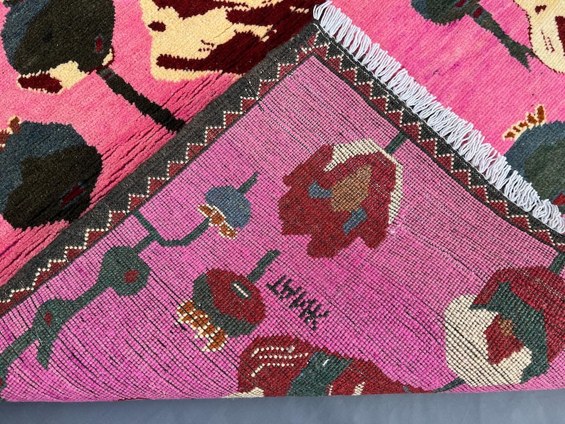 K&ouml;nnte beinhalten: Ein leuchtend rosa Teppich mit einem Blumenmuster in Dunkelgr&uuml;n, Creme und Burgunderrot. Der Teppich hat einen dunkelgrauen Rand mit Zickzackmuster und wei&szlig;en Fransen. Das Design zeigt stilisierte Blumen und geometrische Formen.