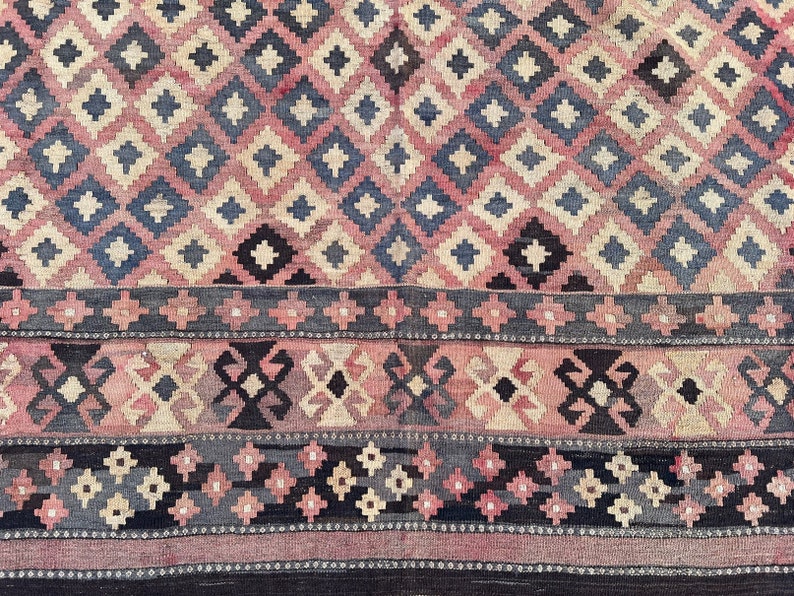 K&ouml;nnte beinhalten: Ein Vintage-Teppich mit einem geometrischen Muster in Rosa-, Blau- und Schwarzt&ouml;nen. Der Teppich hat ein Rautenmuster mit einem Rand aus kleineren geometrischen Formen.