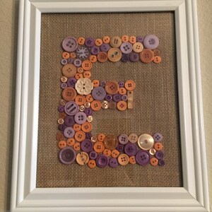 May include: A framed artwork featuring the letter "E" created with a variety of buttons in shades of orange and purple. The buttons are arranged on a burlap background.