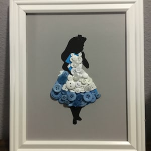 Puede incluir: Una silueta enmarcada de Alicia en el País de las Maravillas hecha con botones azules y blancos. La silueta está sobre un fondo gris.