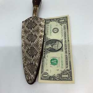 May include: A knife with a snakeskin-patterned sheath, a dark wooden handle, and a bone-colored section. A one-dollar bill is placed next to the knife for size comparison. The sheath is brown and tan.