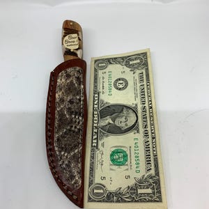 Könnte beinhalten: Ein handgefertigtes Messer mit braunem Griff und einer Scheide mit Schlangenmuster. Das Messer liegt neben einem Ein-Dollar-Schein. Der Griff trägt den Text "Skin Shop". Das Messer wird wahrscheinlich für die Jagd oder Outdoor-Aktivitäten verwendet.