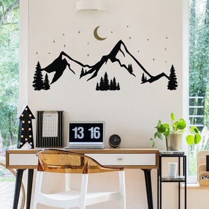 Può includere: Un moderno ufficio domestico con una scrivania bianca, una sedia in plastica trasparente e un laptop che mostra "13 16". Un adesivo da parete nero con montagne e alberi è sopra la scrivania. Sono visibili anche un piccolo orologio nero e una pianta in vaso.