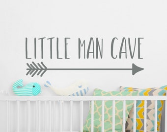 Op de afbeelding: Een witte wieg met een wit matras en decoratieve kussens. Op de muur boven de wieg staat de tekst "LITTLE MAN CAVE" met een pijl. Twee walvisspeeltjes en een gestreept visspeeltje liggen op de wieg.