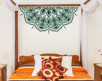 May include: A bedroom with a wooden bed frame and a patterned orange and brown bedspread. Decorative pillows in red, brown, and gold sit on the bed. A green mandala design is on the wall above the bed.