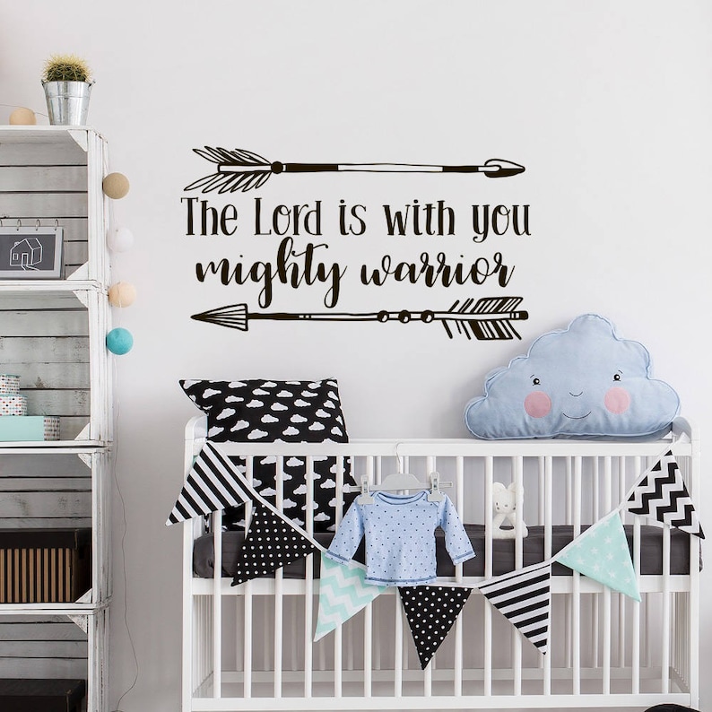 May include: A nursery scene with a white crib decorated with a banner of black and white triangles and a blue cloud-shaped pillow. The wall features a black arrow design and the text "The Lord is with you mighty warrior."