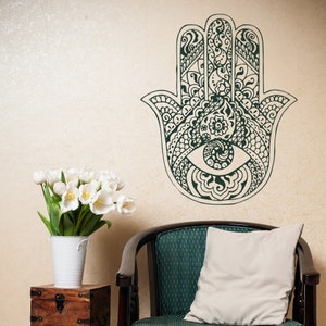 May include: A black and white hamsa hand wall decal with intricate details and an eye in the center. The decal is on a beige wall above a teal armchair with a white pillow.