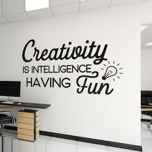 May include: A white office wall with black text that reads "Creativity is intelligence having fun" and a lightbulb graphic. A desk with a computer and chair is in the foreground. The office has a modern design.