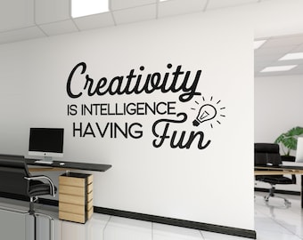 May include: A white office wall with black text that reads "Creativity is intelligence having fun" and a lightbulb graphic. A desk with a computer and chair is in the foreground. The office has a modern design.